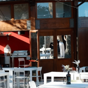 Outdoor patio of a restaurant with white tables and chairs, a red wall, and wooden doors reflecting a person inside.