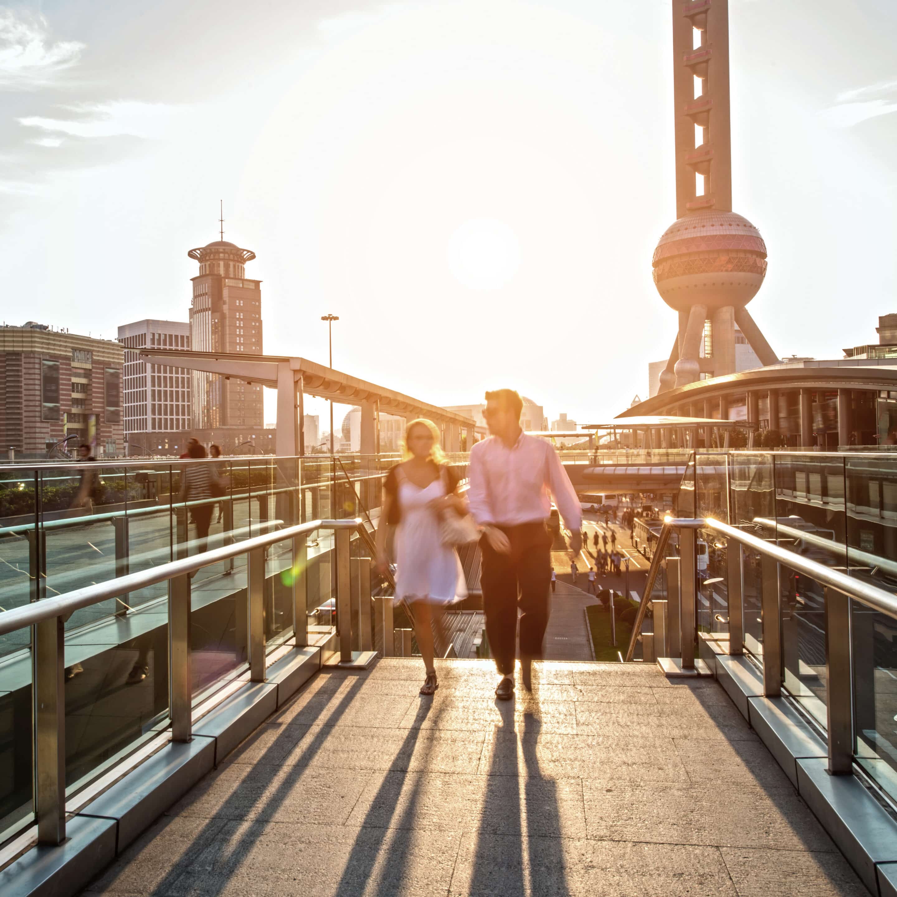 Couple walks on an elevated pedestrian bridge in Shanghai with the Oriental Pearl TV Tower and city skyline under bright sun.
