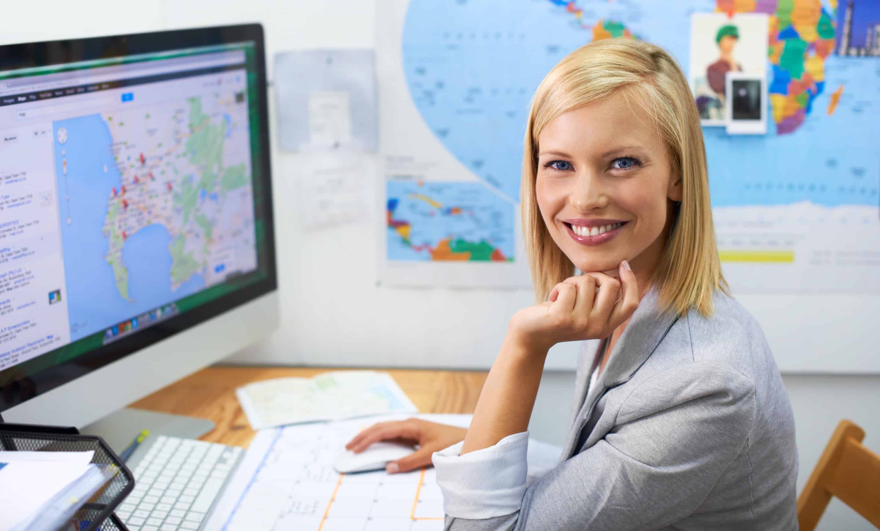 Smiling woman working at a computer with a map-based tourism app on screen, with a world map behind her.