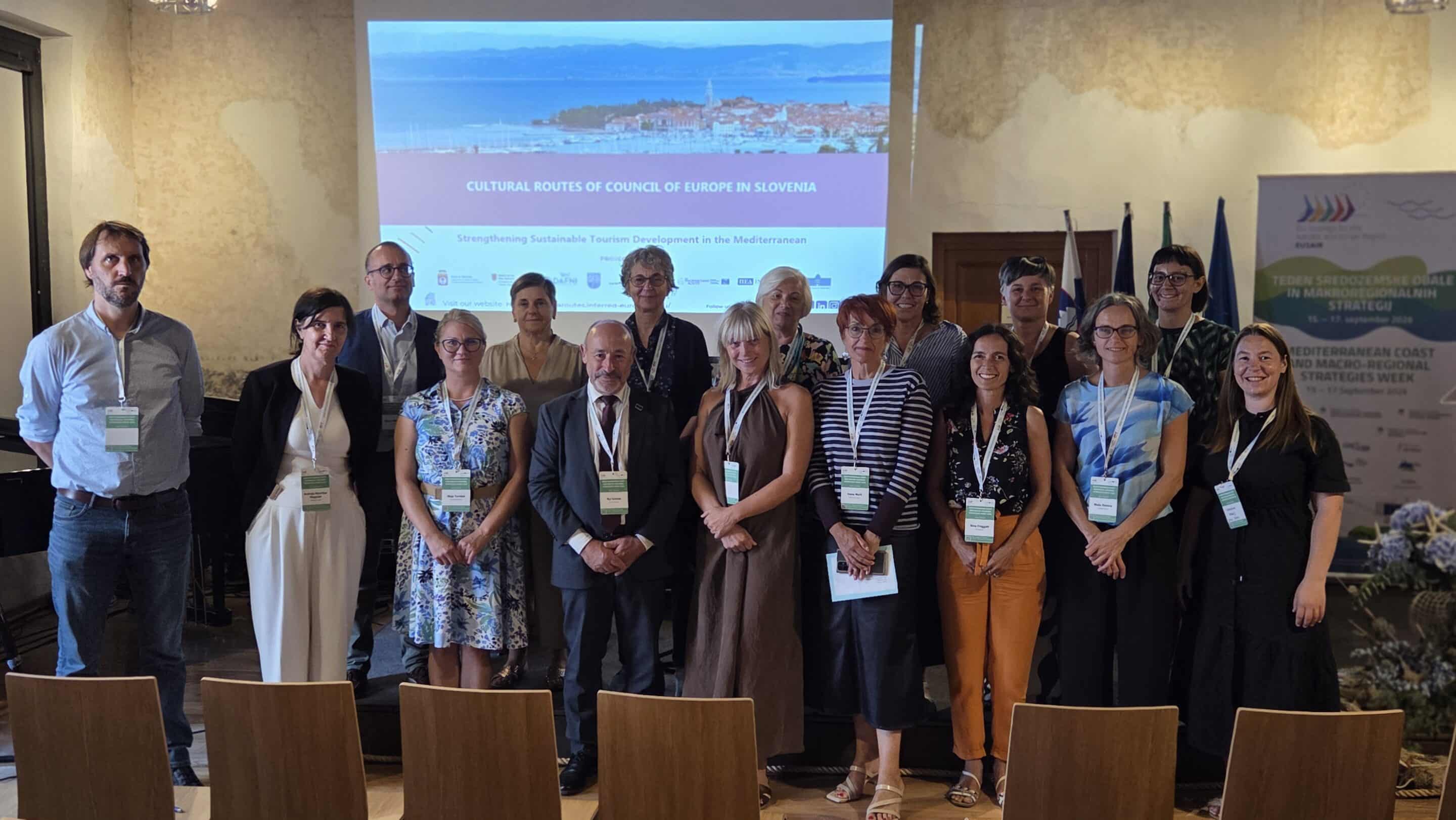 Group of 17 participants, mostly women, smiling in front of a screen showing 'Cultural Routes of Council of Europe in Slovenia' and a coastal town.