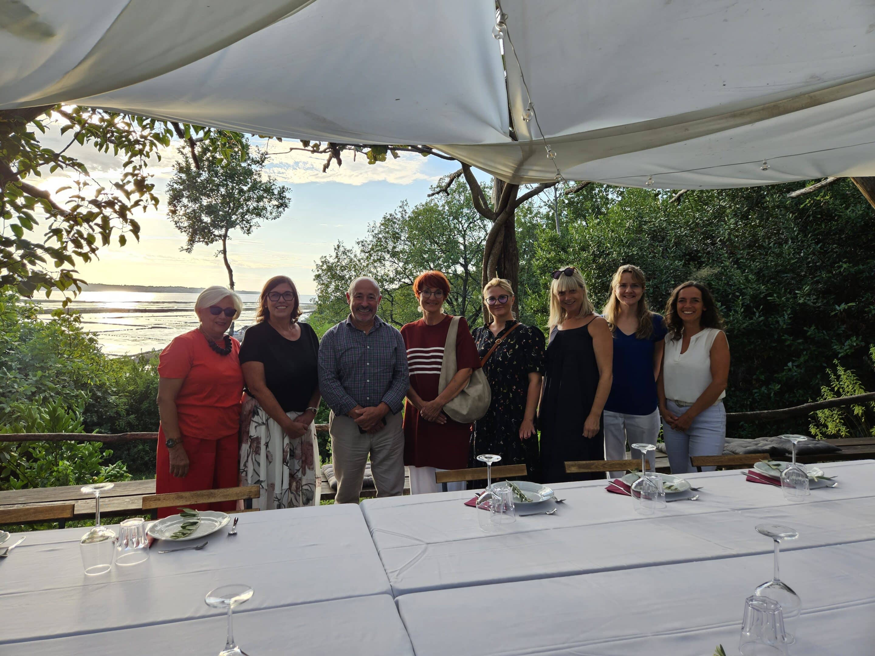 Eight people, two men and six women, stand behind a long white table under a canopy, overlooking a scenic bay and trees at sunset.